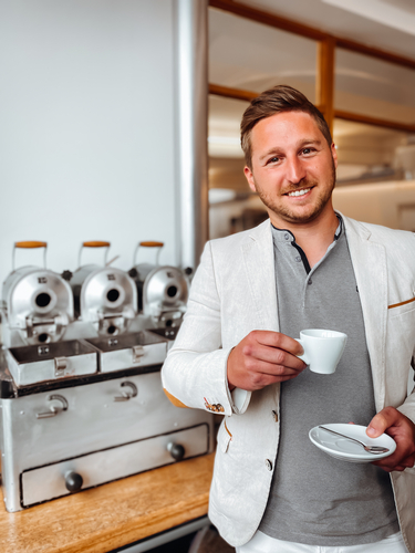 Office-Barista Stefan Riegler blickt lächelnd in die Kamera und hält eine Tasse Kaffee und eine Untertasse, im Hintergrund alte Barista-Ausrüstung