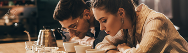 Eine junge Frau und ein junger Mann beugen sich über mehrere Kaffeetassen vor ihnen auf dem Tisch und riechen daran