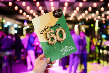 Eine Glückwunschkarte mit Aufschrift "Unglaubliche 50", im Hintergrund Partylichter