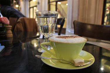 Eine Tasse Kaffee mit Milchschaum und ein Wasserglas, im Hintergrund das Innere eines Cafés