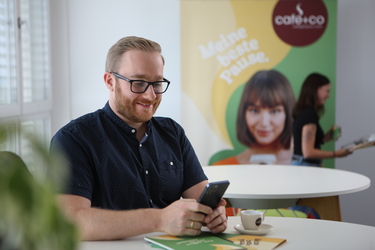 Ein Mann sitzt an einem runden Tisch mit einer Kaffeetasse und blickt lächelnd auf sein Smartphone