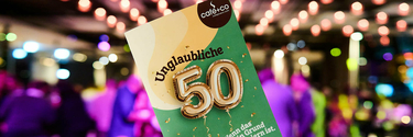 Eine Glückwunschkarte mit Aufschrift "Unglaubliche 50", im Hintergrund Partylichter