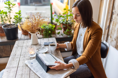 Junge Frau sitzt an einem Holztisch und blickt auf ihr Tablet, daneben eine Tasse Kaffee, Zeitung und Smartphone, im Hintergrund Topfpflanzen