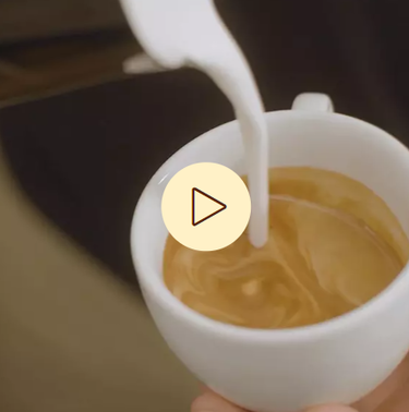 Nahaufnahme einer Tasse Kaffee beim Einschenken von Milch, im Vordergrund ein Symbol zum Abspielen von Videos