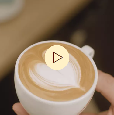 Nahaufnahme einer Tasse Kaffee mit Milchschaum, im Vordergrund ein Symbol zum Abspielen von Videos