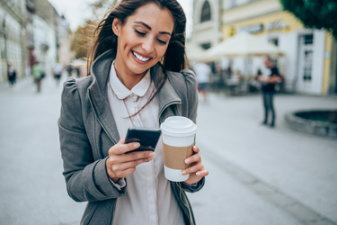 Junge Frau geht auf der Straße mit einem Kaffeebecher und blickt lachend auf ihr Smartphone