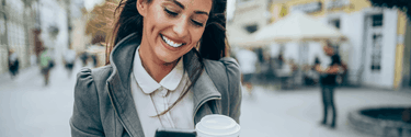 Junge Frau geht auf der Straße mit einem Kaffeebecher und blickt lachend auf ihr Smartphone